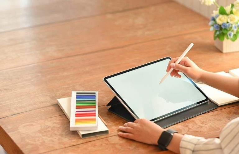 Eine Hand zeichnet mit einem Stylus auf einem Tablet, neben dem Farbstifte liegen.